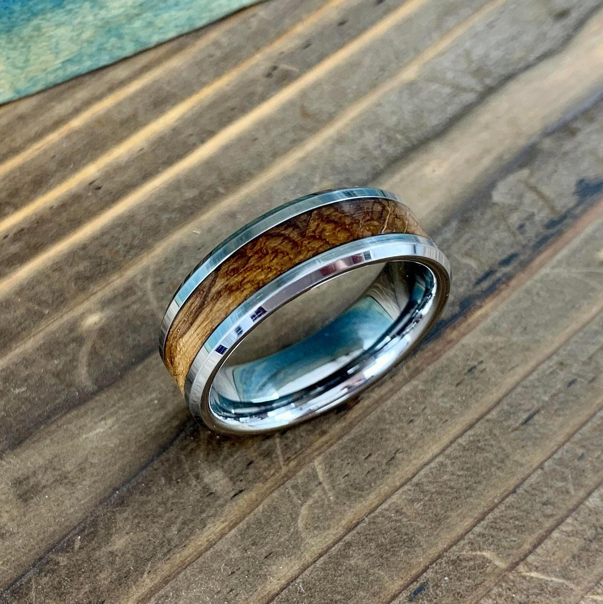 A silver-colored ring with a central light brown wood band, on a wooden surface with a blue-green background.