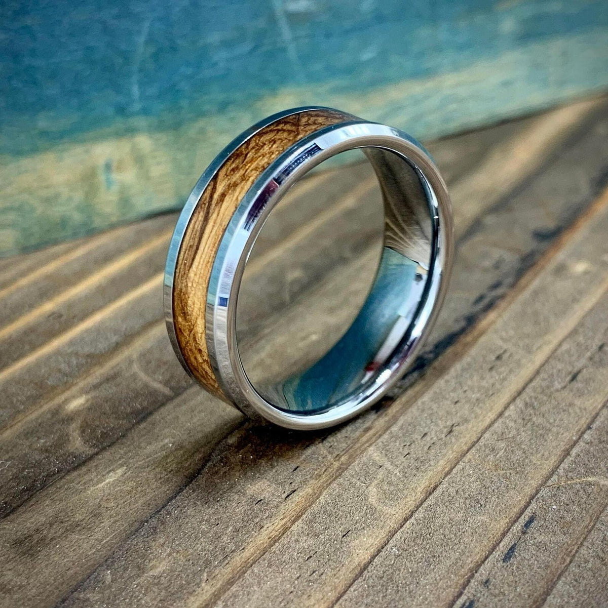 A silver-colored ring with a light brown wood inlay, on a wooden surface with a blue-green background.