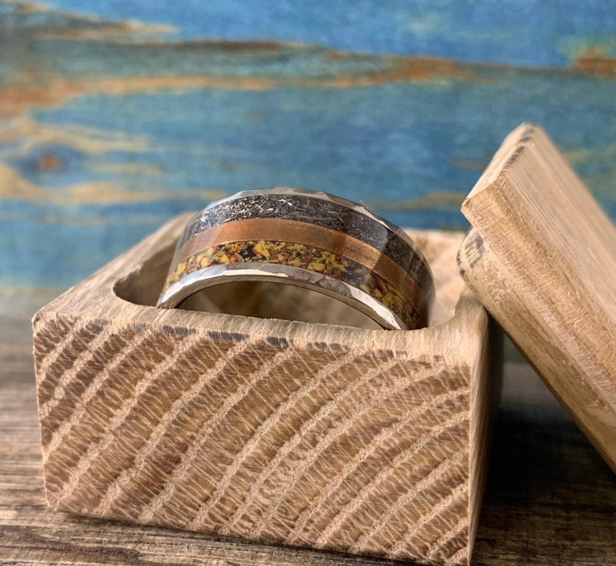 A silver-colored ring with hammered outer edges, a smooth copper-colored band, and a textured yellow-orange band in the center, resting in an open wooden box.