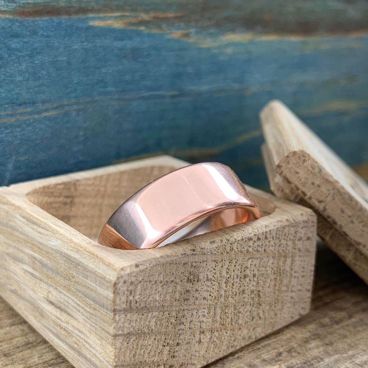 A matte rose gold-colored ring with a slightly rounded top surface rests inside an open light wood box.