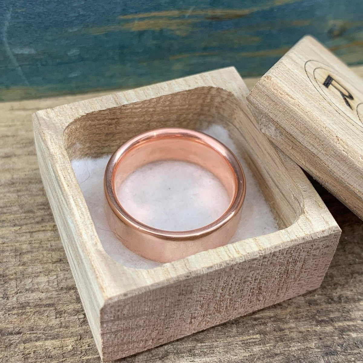 A shiny rose gold-colored ring with a flat top surface rests inside an open light wood box, viewed from above.