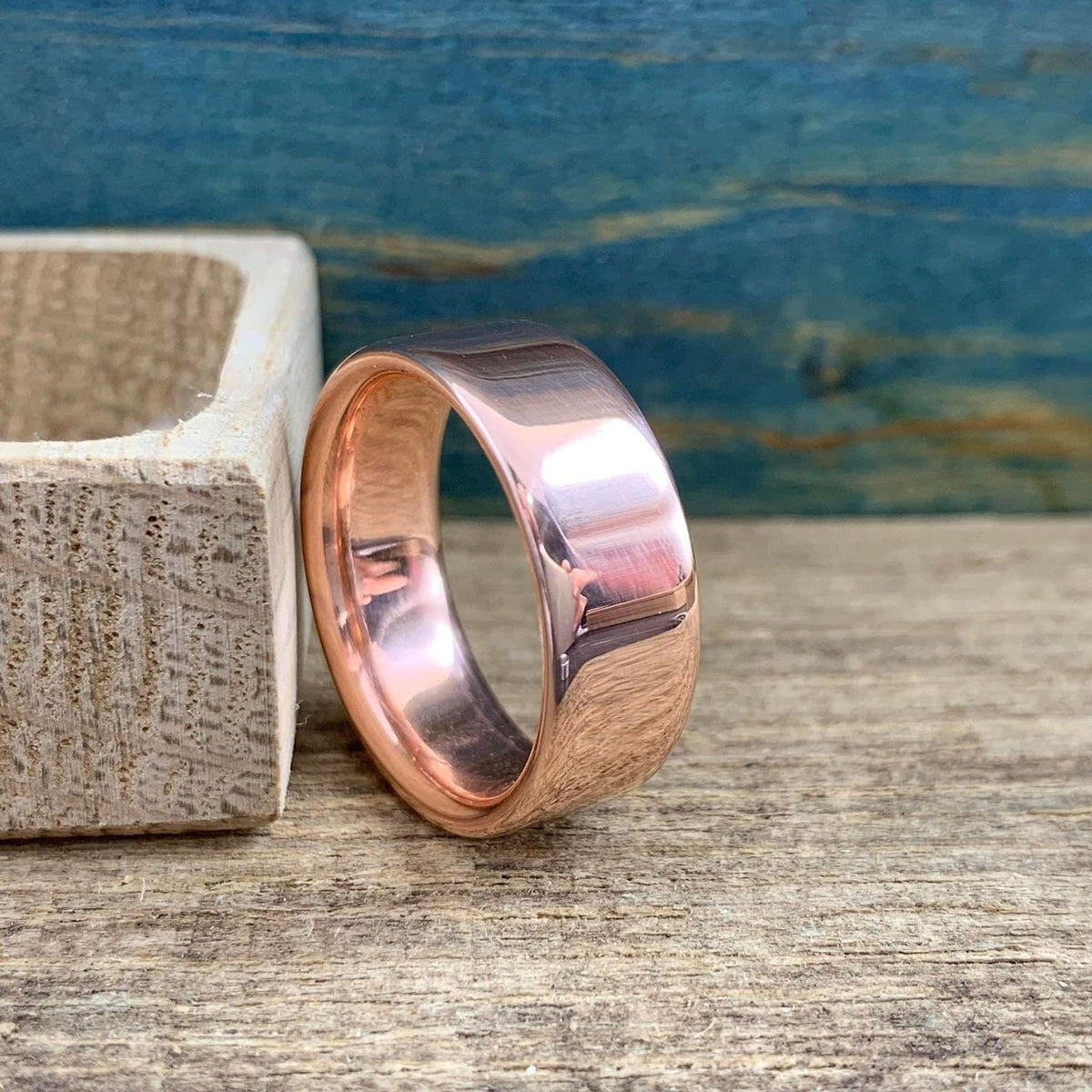 A shiny rose gold-colored ring with a slightly rounded top surface is next to an open light wood box.