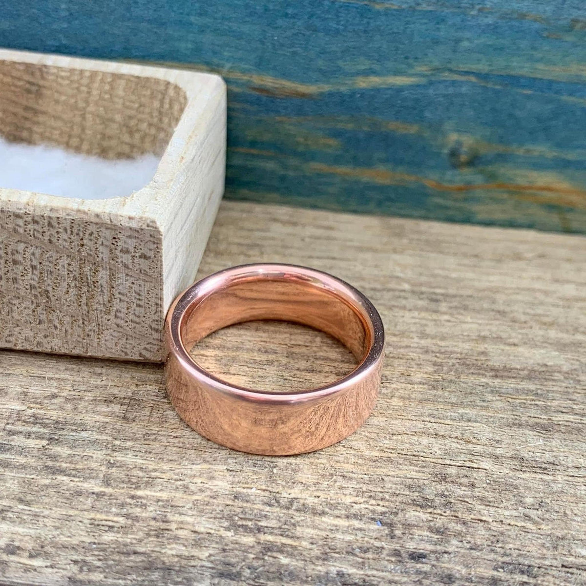 A shiny rose gold-colored ring with a flat top surface is next to an open light wood box, showing the inside of the ring.