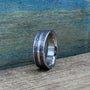 A silver-colored ring with two textured dark gray bands separated by a thin silver-colored stripe, on a wooden surface.