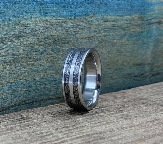 A silver-colored ring with two textured dark gray bands separated by a thin silver-colored stripe, on a wooden surface.