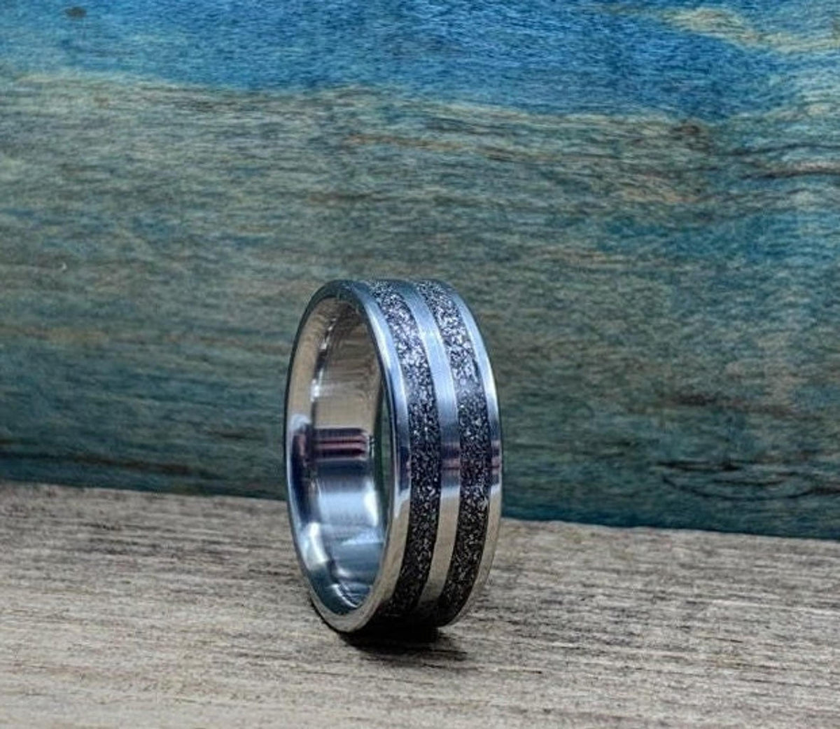A silver-colored ring with two textured dark gray bands separated by a thin silver-colored stripe, on a wooden surface, viewed from a slightly different angle.