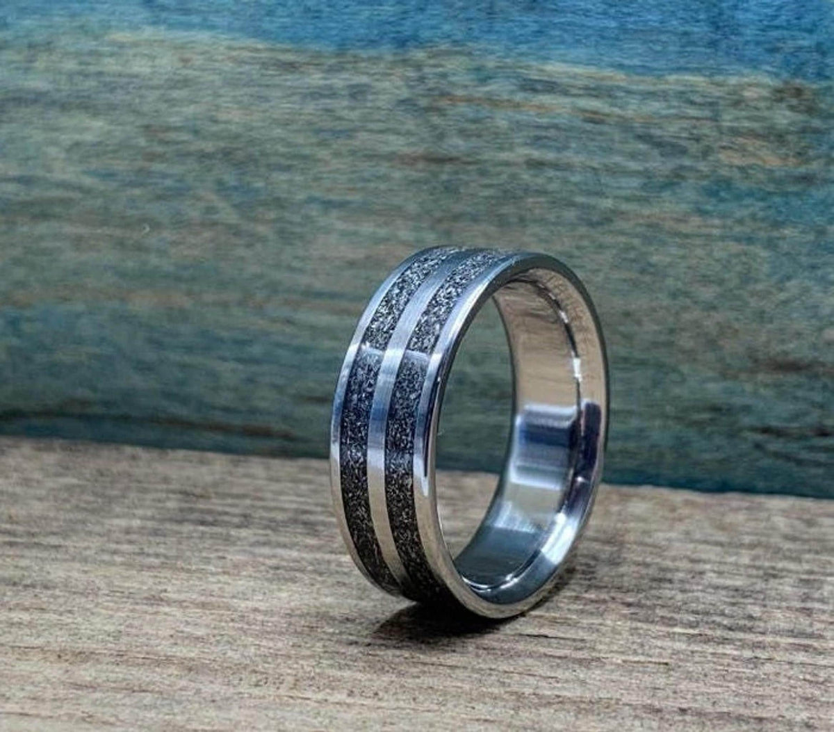 A silver-colored ring with two textured dark gray bands separated by a thin silver-colored stripe, on a wooden surface, viewed from a slightly different angle.
