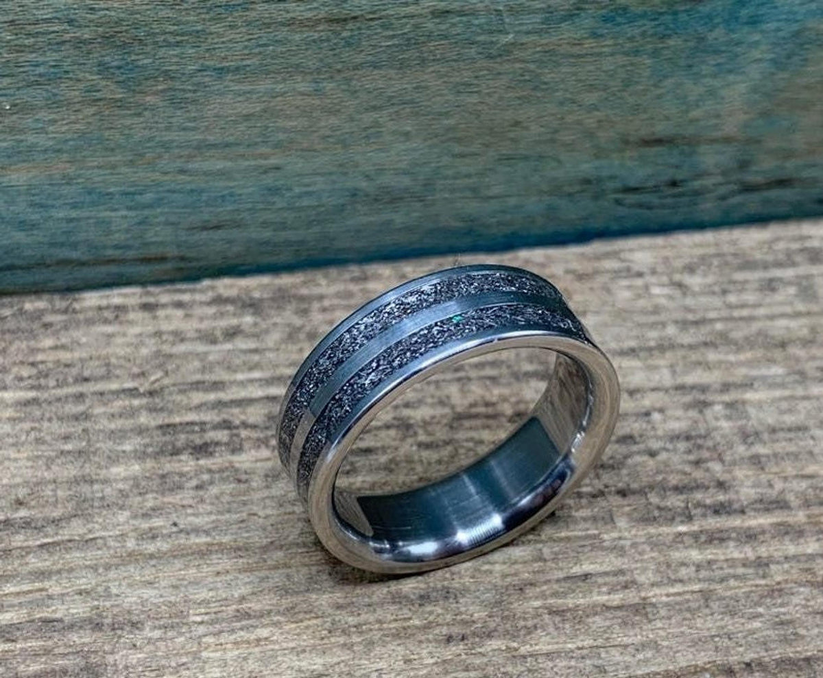 A silver-colored ring with two textured dark gray bands separated by a thin silver-colored stripe, on a wooden surface, viewed from a slightly different angle.