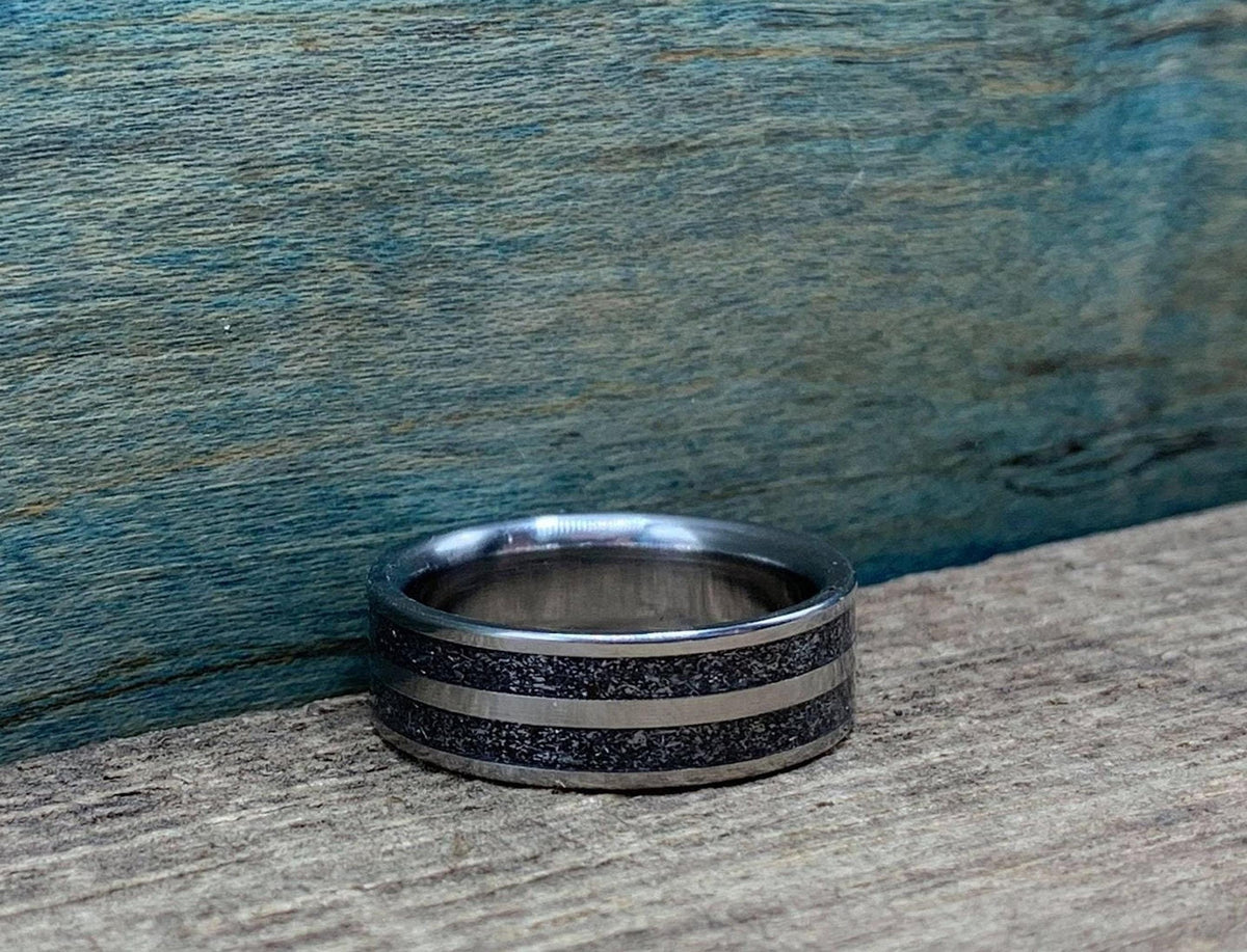 A silver-colored ring with a wide textured dark gray band in the center, flanked by two thin silver-colored stripes, on a wooden surface.