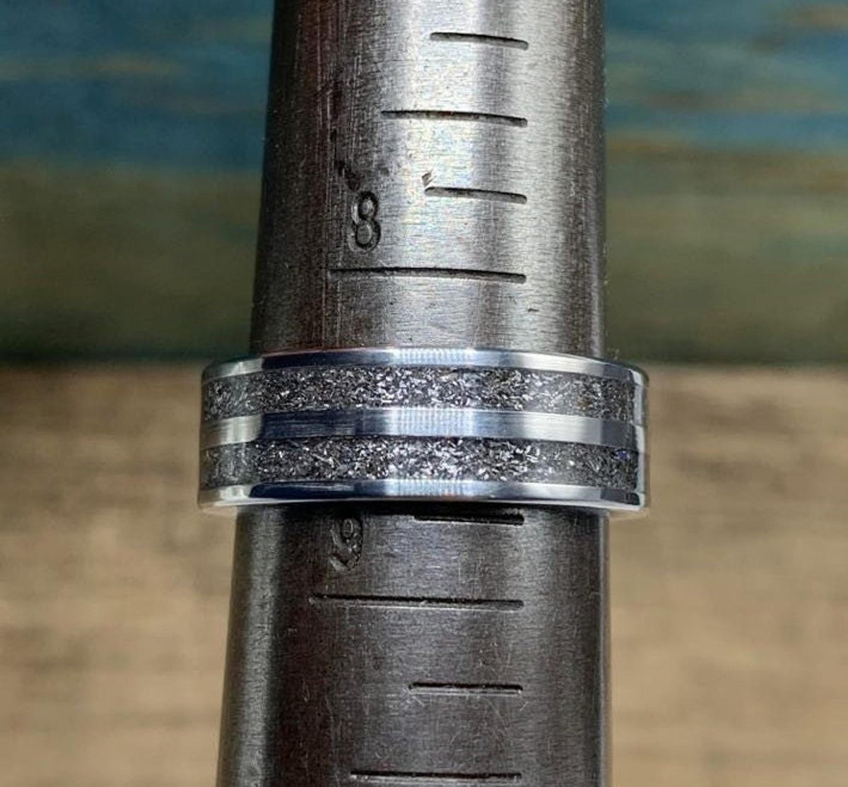 A silver-colored ring with a wide textured dark gray band in the center, flanked by two thin silver-colored stripes, placed on a metal ring sizer showing size 8.
