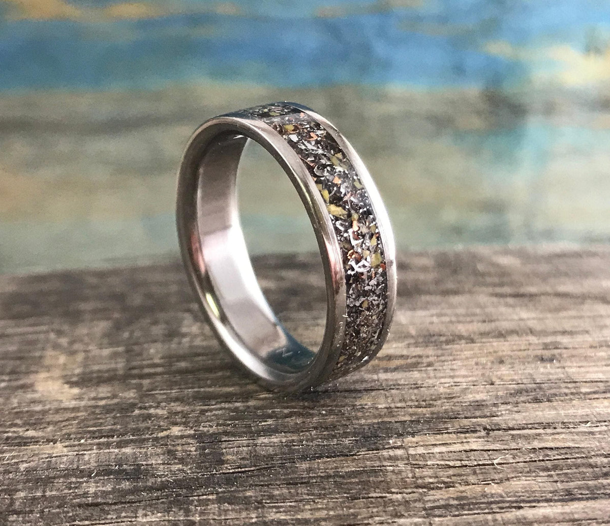 A silver-colored ring with a central band of speckled, multi-colored material is angled on a weathered wooden surface.