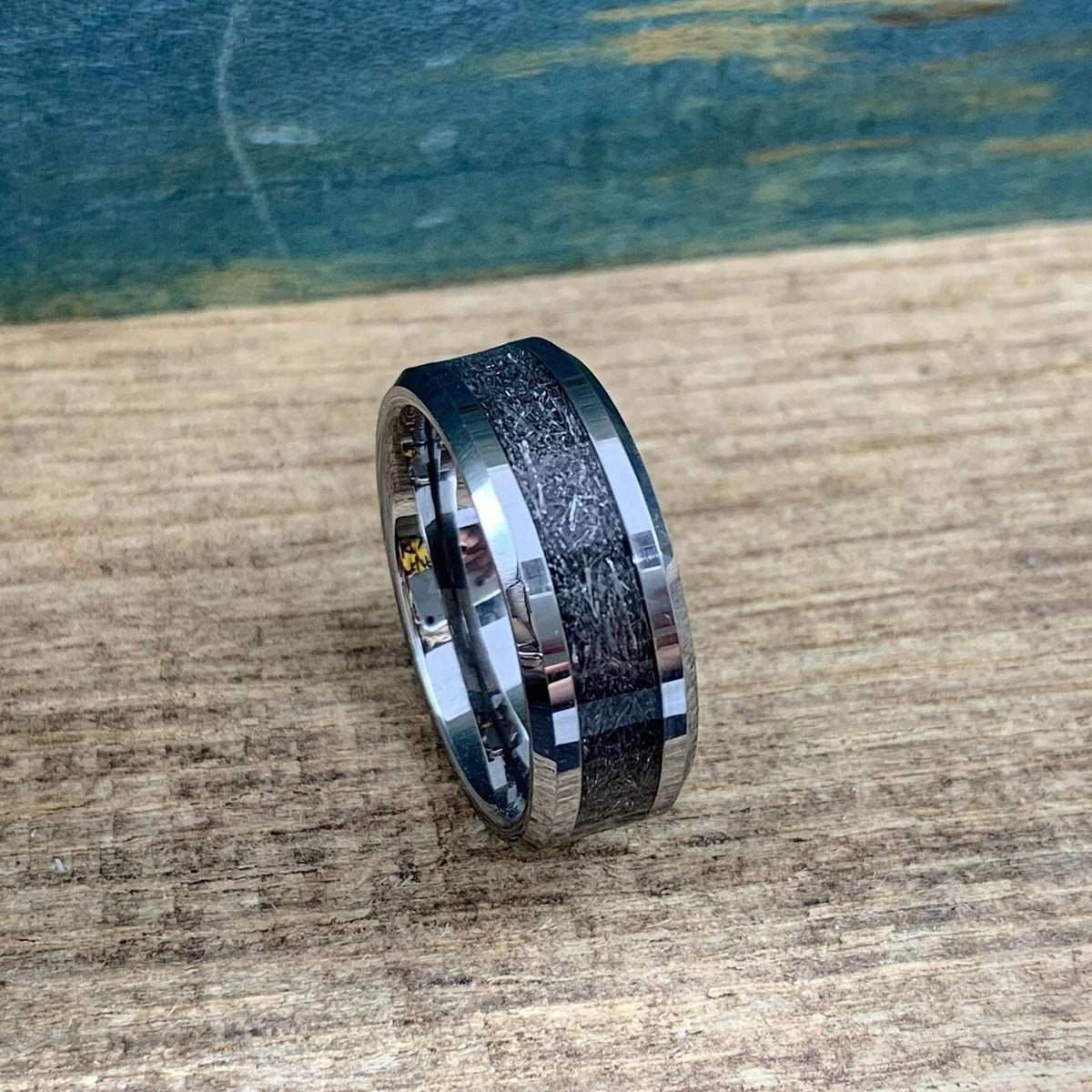 A silver-toned tungsten ring with a dark, textured meteorite inlay is angled on a wooden surface, showing the side and top.