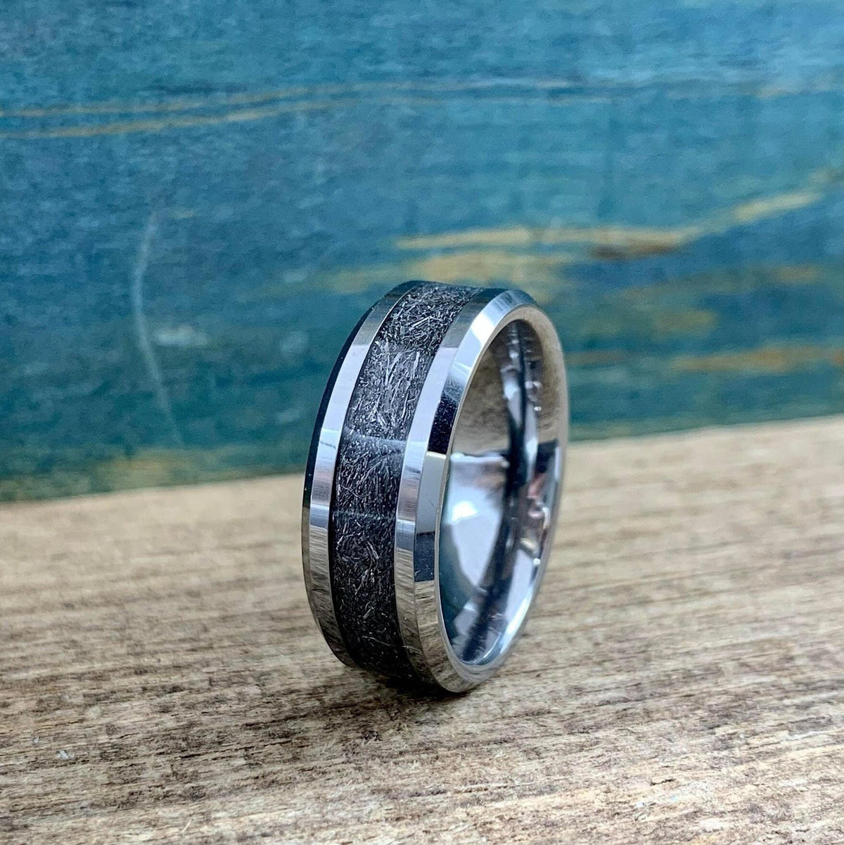 A silver-toned tungsten ring with a dark, textured meteorite inlay is shown from a slightly different angle on a wooden surface.