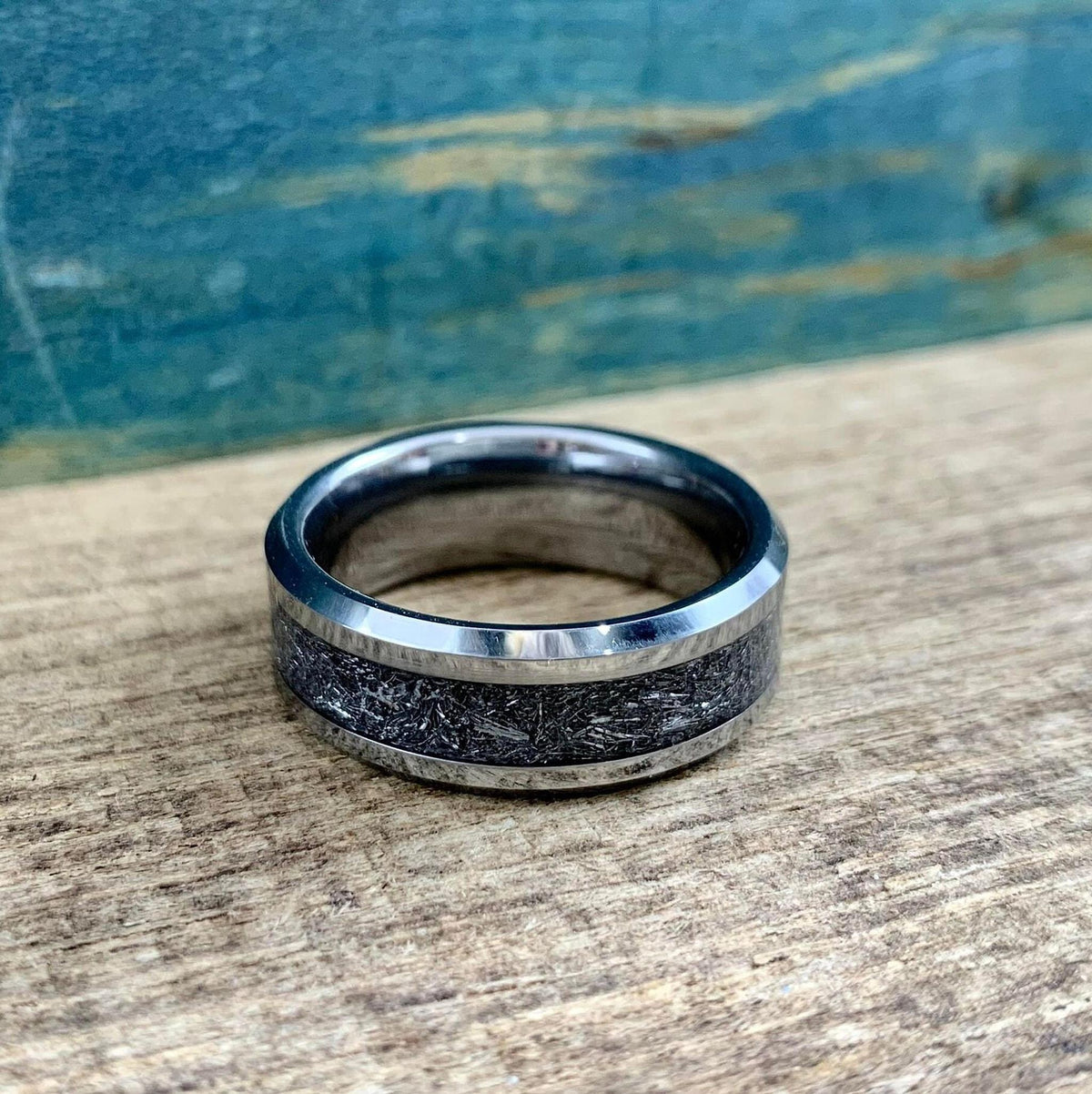 A silver-toned tungsten ring with a dark, textured meteorite inlay is shown from a top-down perspective on a wooden surface.