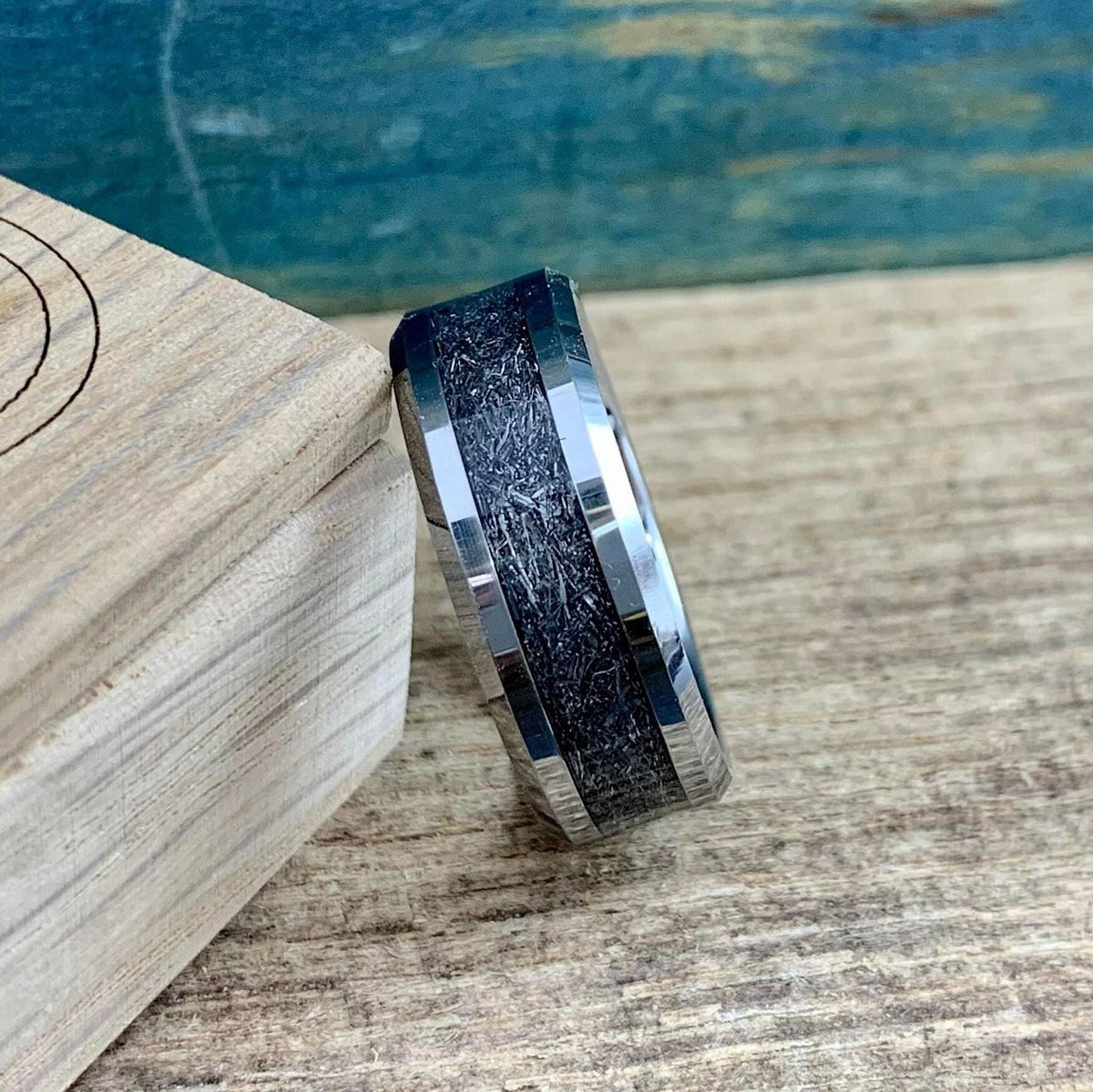 A silver-toned tungsten ring with a dark, textured meteorite inlay is propped against a light wooden block.