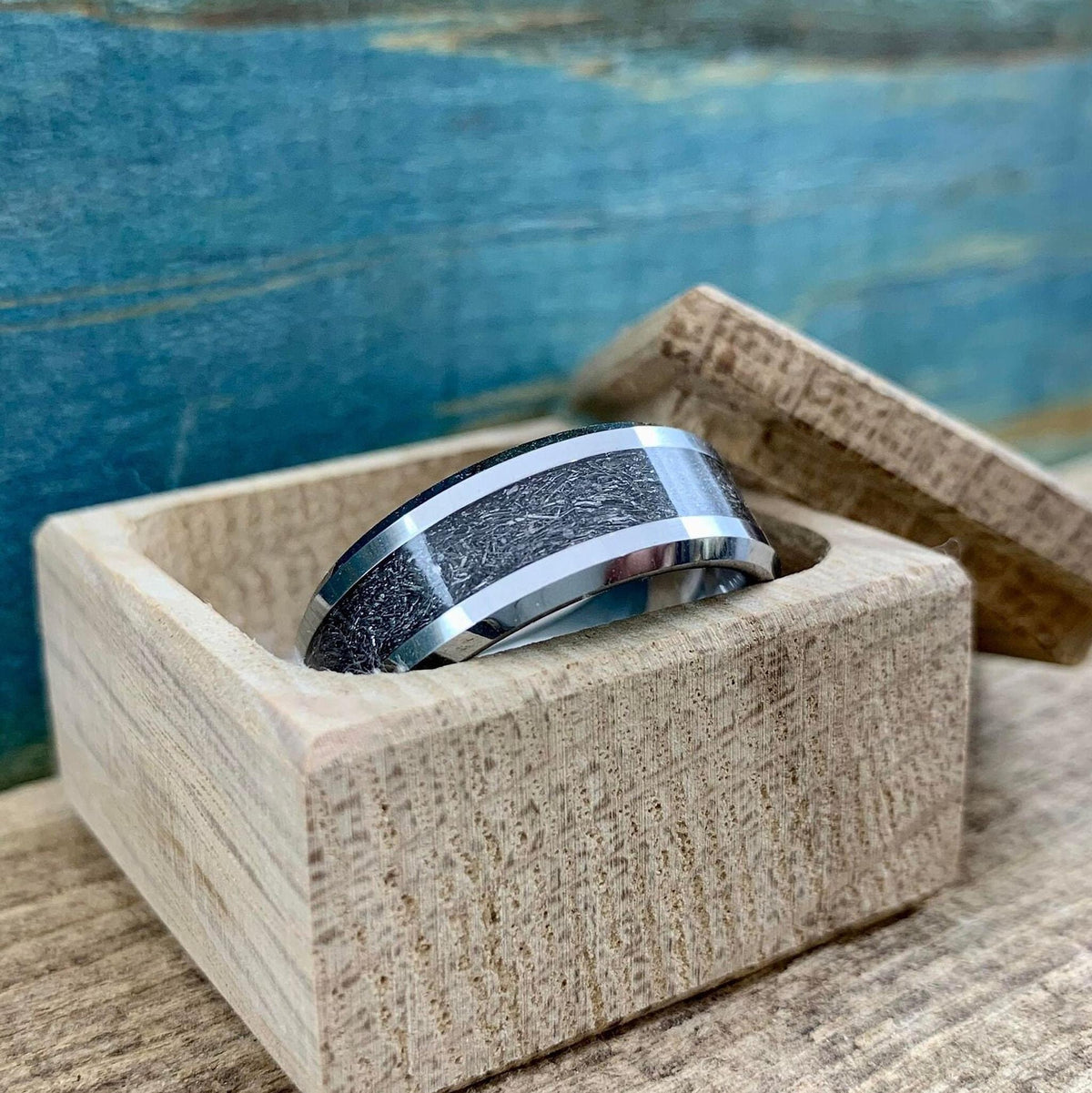 A silver-toned tungsten ring with a dark, textured meteorite inlay rests inside a small, light wooden box.