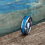 A silver-colored ring with a central blue-green opal inlay is angled on a wooden surface with a blue background.