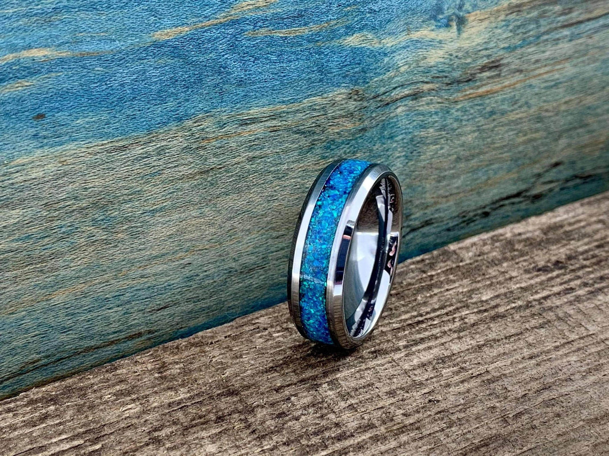 A silver-colored ring with a central blue-green opal inlay is angled on a wooden surface with a blue background.