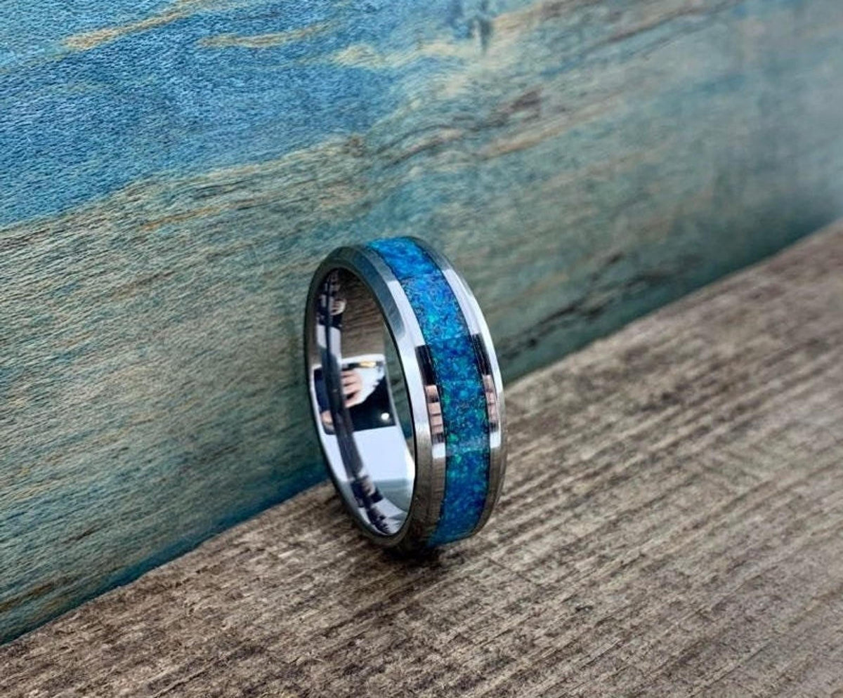 A silver-colored ring with a blue-green opal inlay and a thin copper-colored line is angled on a wooden surface.