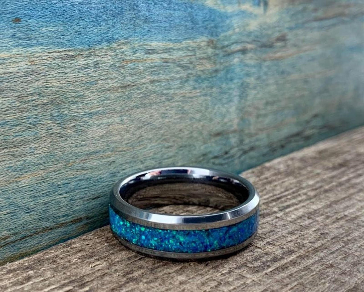 A silver-colored ring with a wide blue-green opal inlay is centered on a wooden surface.