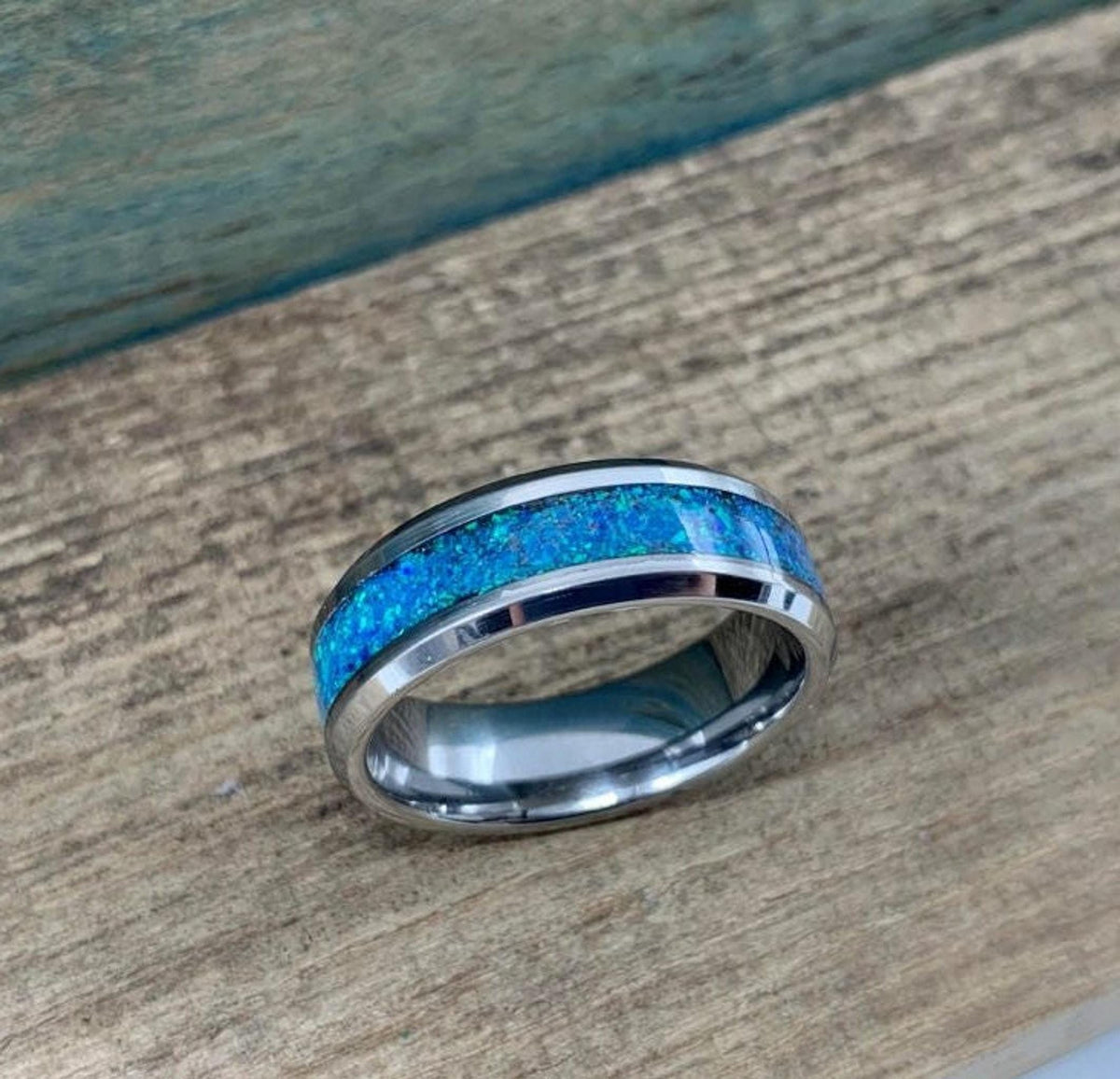 A silver-colored ring with a blue-green opal inlay is shown on a light brown wooden surface.