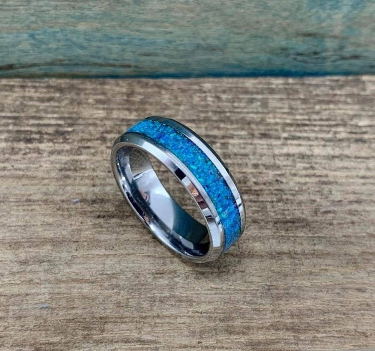 A silver-colored ring with a blue-green opal inlay is shown from a top-down angle on a wooden surface.