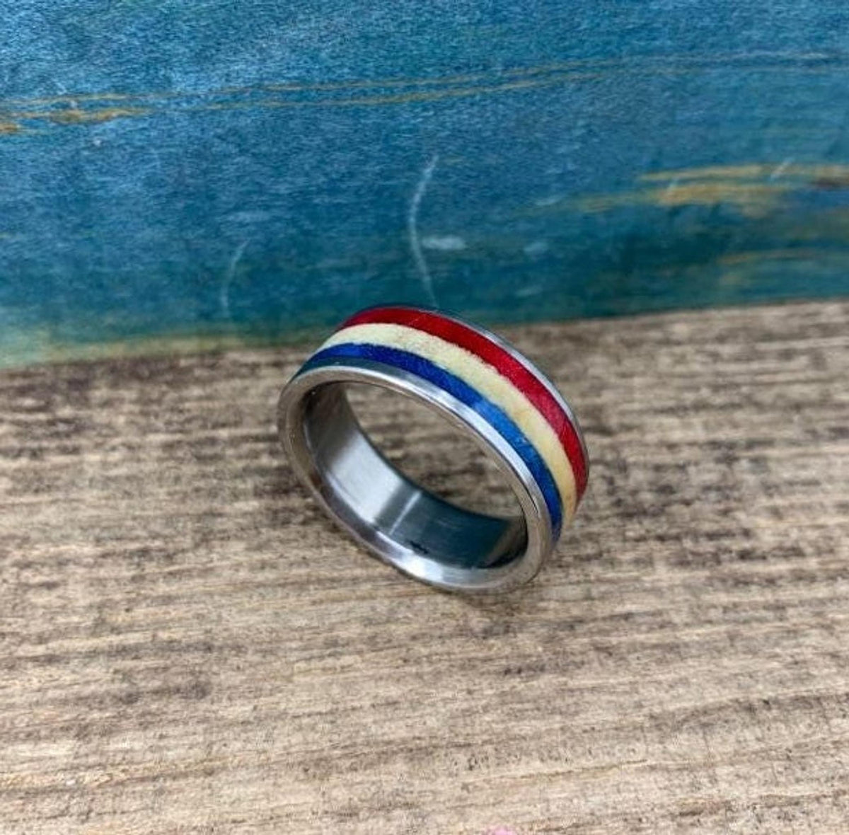 A silver-colored ring with red, white, and blue stripes is shown from a slightly elevated angle, slightly to the left, on a wooden surface.