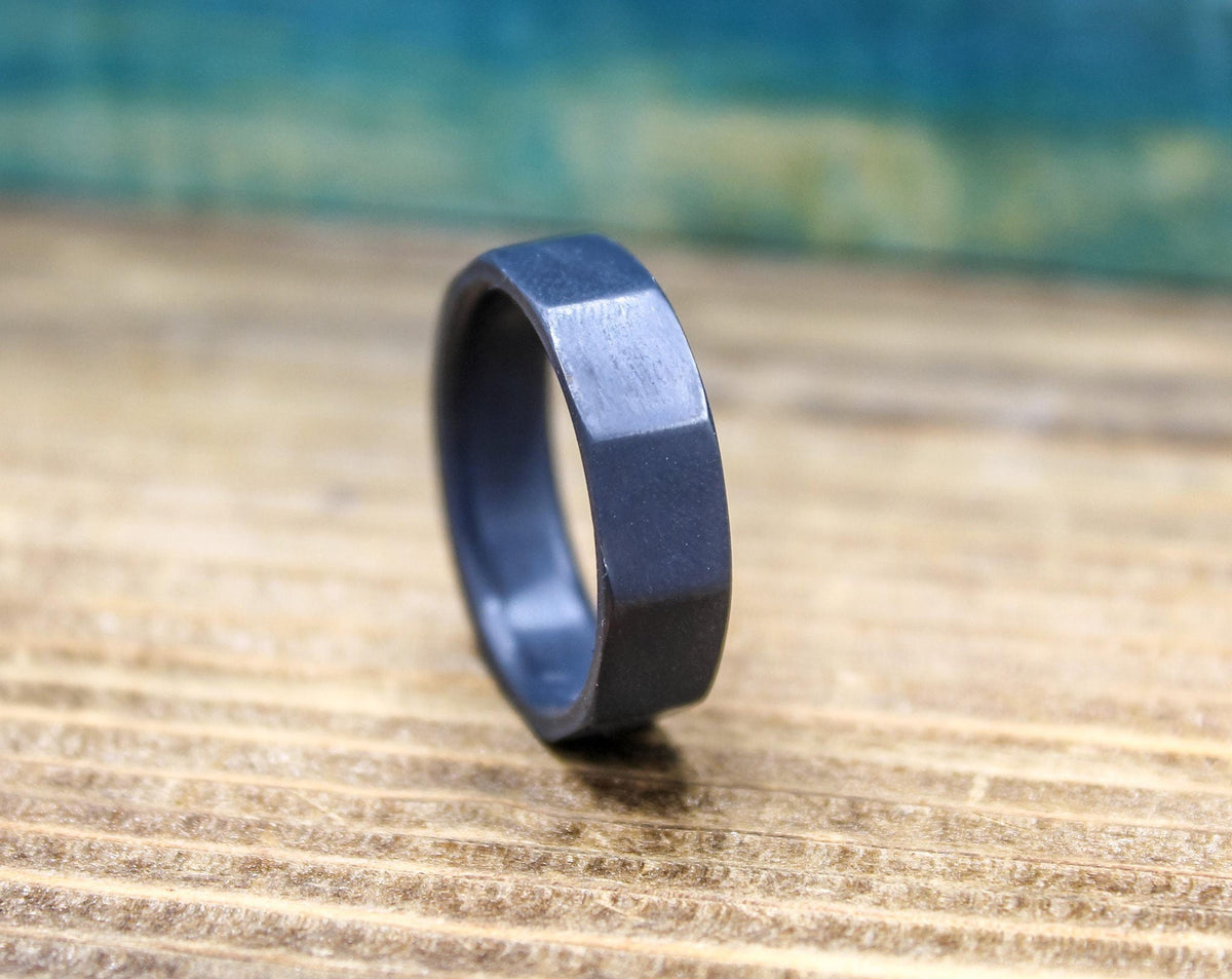 A dark gray, ten-sided ring lies flat on a light brown wooden surface, with a blurred blue background.