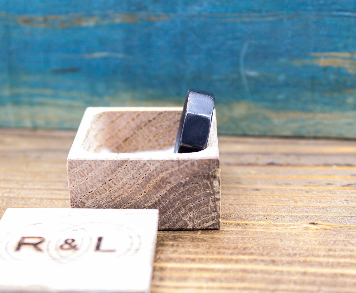 A dark gray, ten-sided ring is visible inside an open light wooden box, with the box lid showing "R & L" in the foreground, and a blurred blue background.