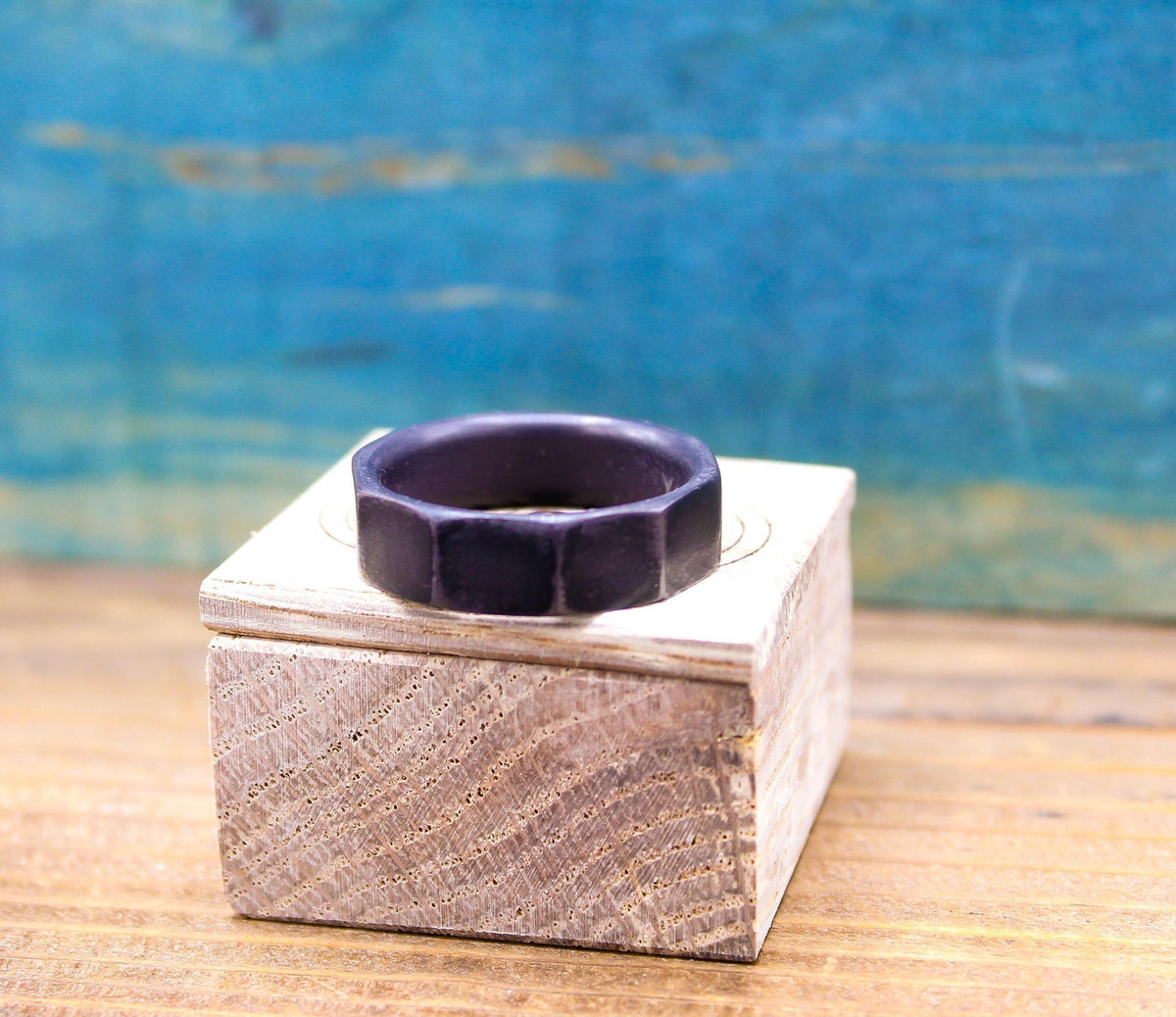 A dark gray, ten-sided ring rests on top of a closed light wooden box, with a blurred blue background.