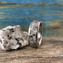 A silver-colored ring with a speckled gray and white granite inlay sits on a wooden surface next to a matching granite rock.