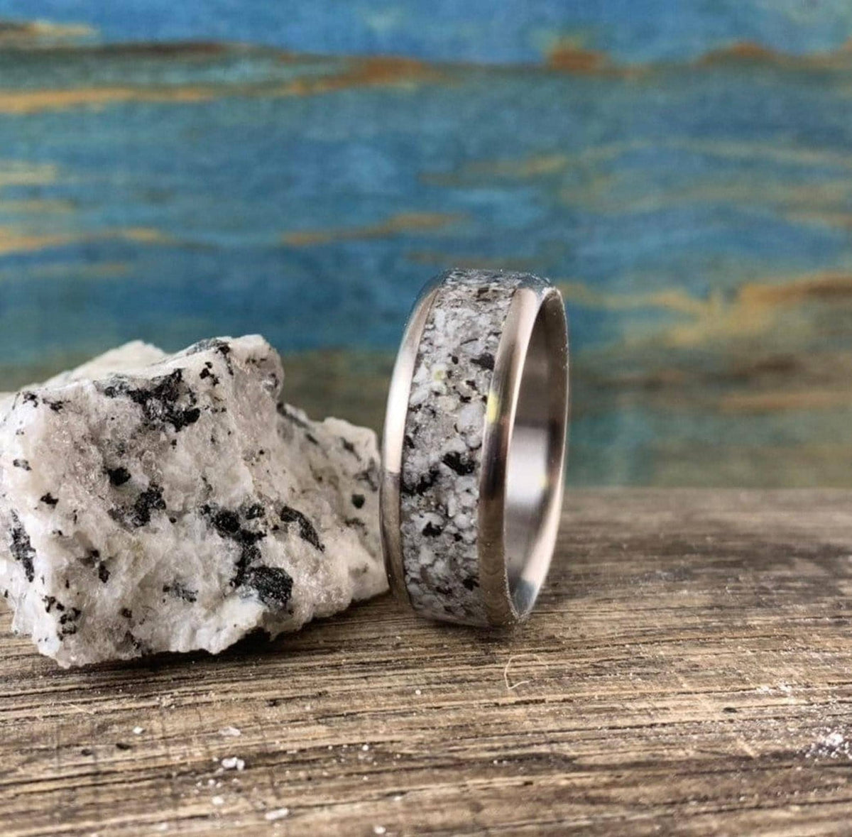 A silver-colored ring with a speckled gray and white granite inlay sits on a wooden surface next to a matching granite rock.