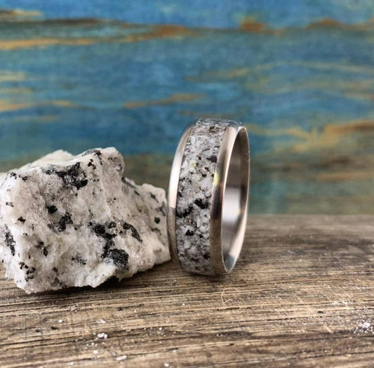 A silver-colored ring with a speckled gray and white granite inlay sits on a wooden surface next to a matching granite rock.