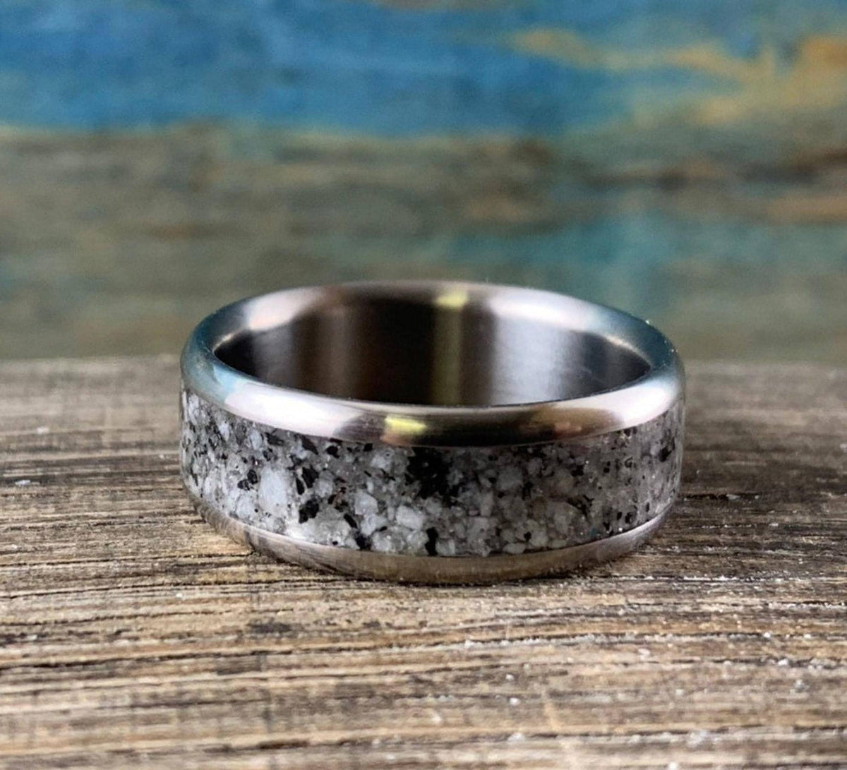 A silver-colored ring with a speckled gray and white granite inlay is centered on a light brown wooden surface.