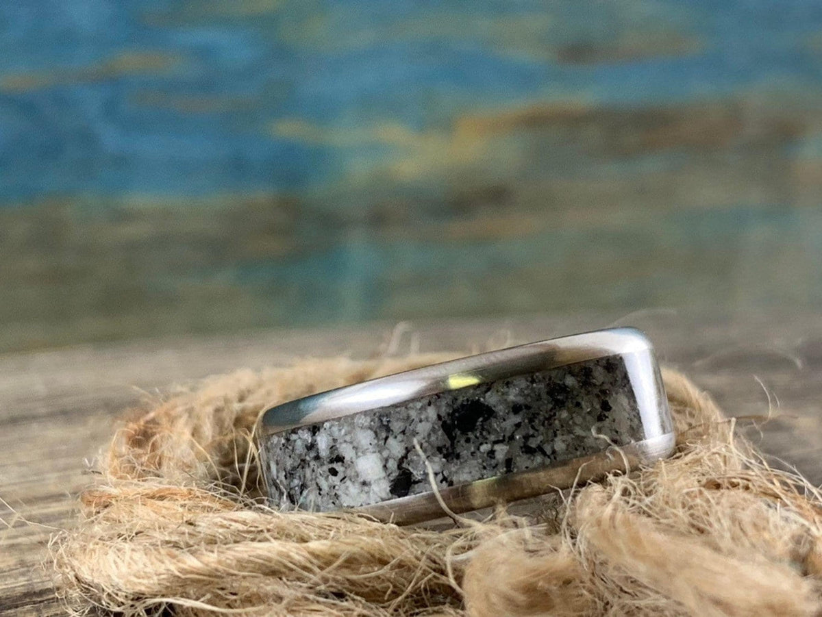 A silver-colored ring with a speckled gray and white granite inlay rests on a bed of natural twine.
