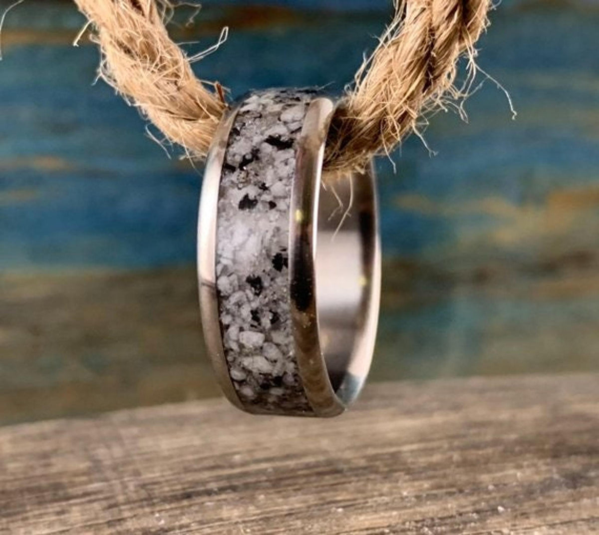 A silver-colored ring with a speckled gray and white granite inlay hangs from a thick piece of natural twine.