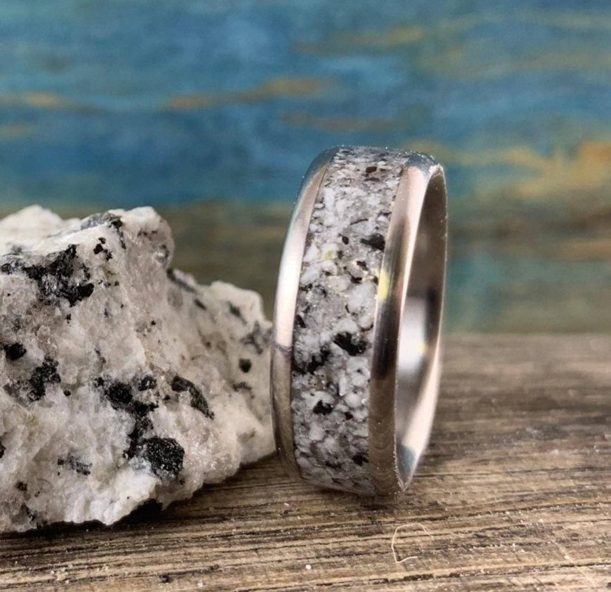 A silver-colored ring with a speckled gray and white granite inlay stands on a wooden surface beside a piece of granite.