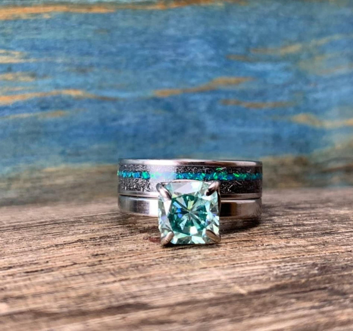 A wider silver-toned band with a black and blue-green inlay sits behind a silver-toned ring with a green square-cut stone, on a wooden surface.