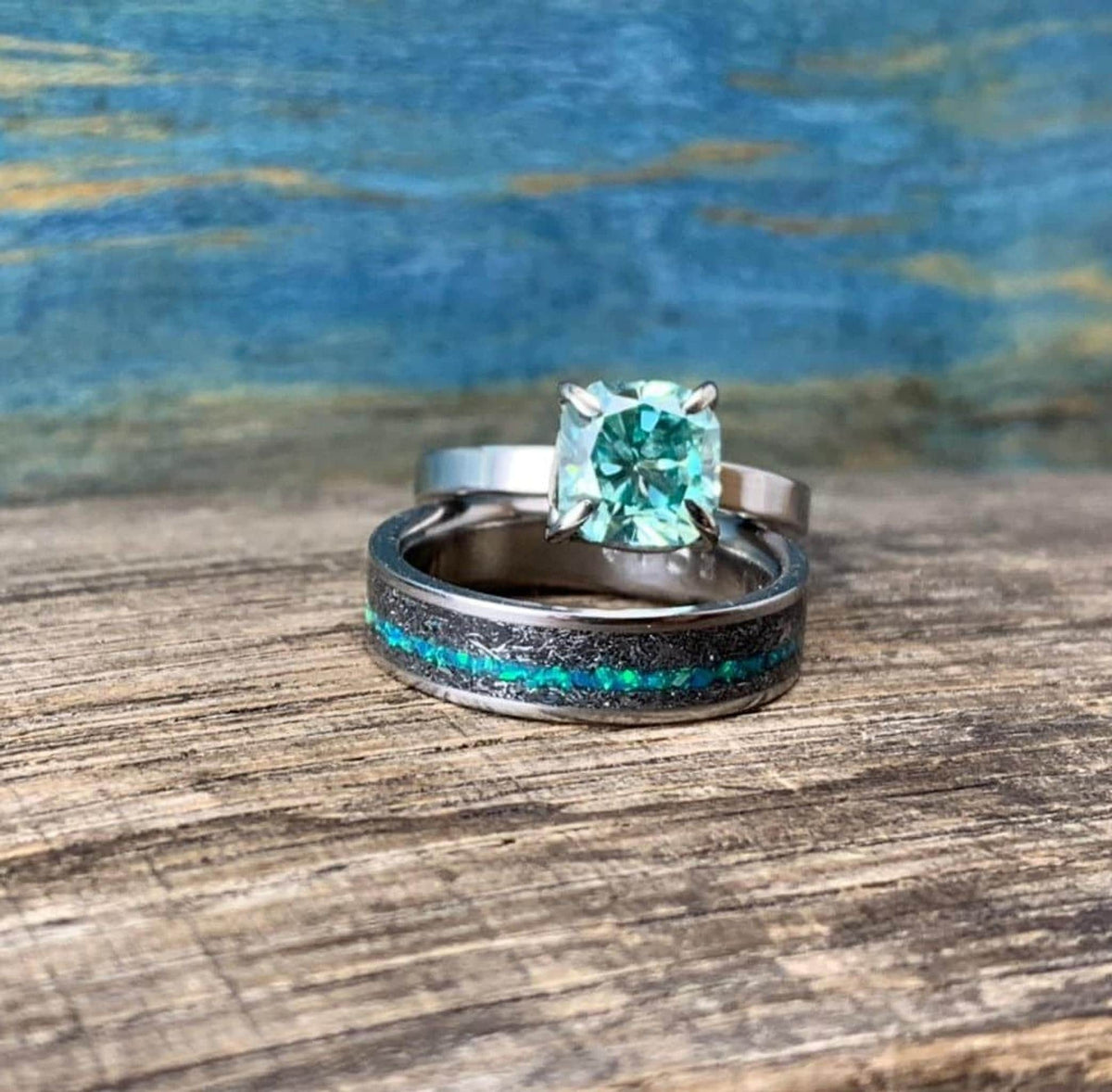 A silver-toned ring with a green square-cut stone is stacked on a wider silver-toned band with a black and blue-green inlay, on a wooden surface.