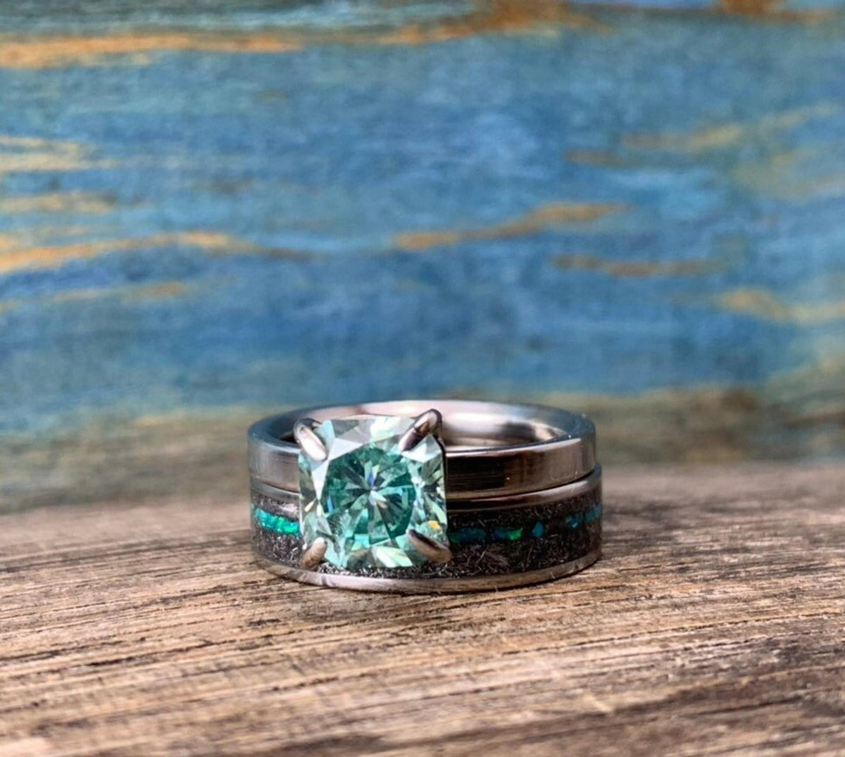 A silver-toned ring with a green square-cut stone is stacked on a wider silver-toned band with a black and blue-green inlay, on a wooden surface.