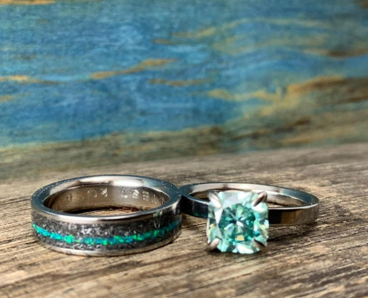 A silver-toned ring with a green square-cut stone sits next to a wider silver-toned band with a black and blue-green inlay, on a wooden surface.