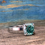 A silver-toned ring with a cushion-cut blue-green stone in a four-prong setting, resting on a wooden surface.