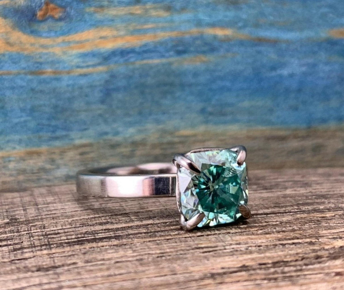 A silver-toned ring with a cushion-cut blue-green stone in a four-prong setting, resting on a wooden surface.