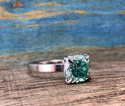 A silver-toned ring with a cushion-cut blue-green stone in a four-prong setting, resting on a wooden surface.