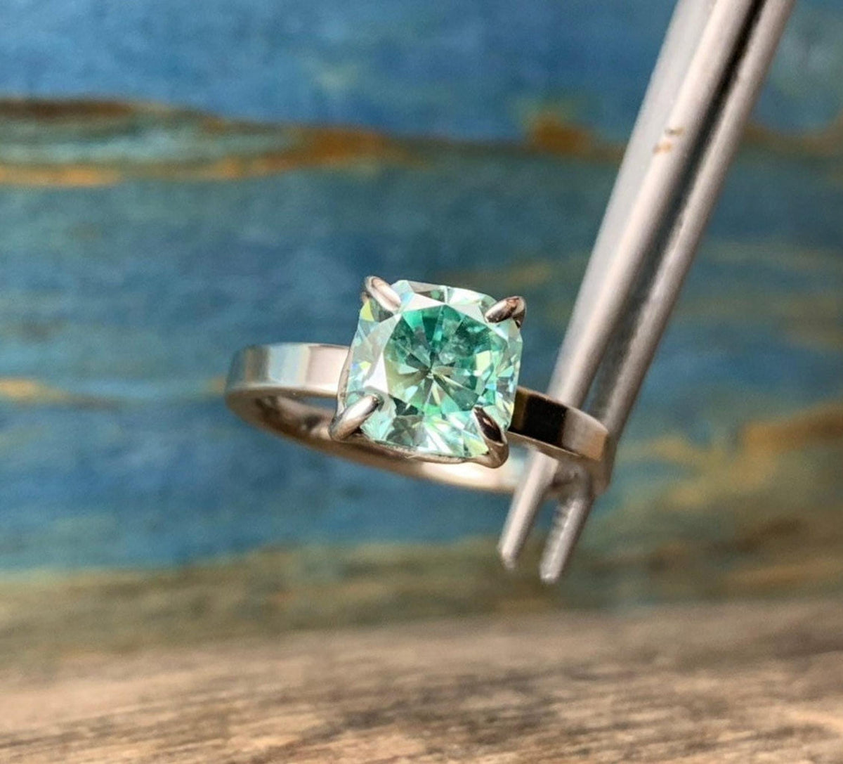 A silver-toned ring with a cushion-cut blue-green stone held by metal tweezers against a wooden surface and blue background.