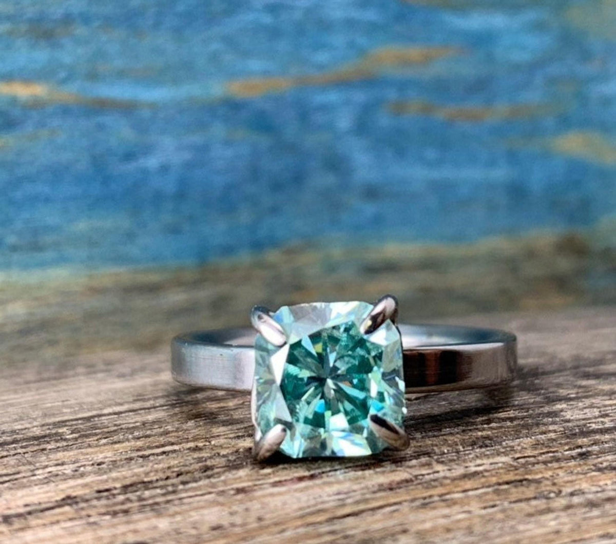 A silver-toned ring with a cushion-cut blue-green stone, centered on a wooden surface with a blurred blue and brown background.