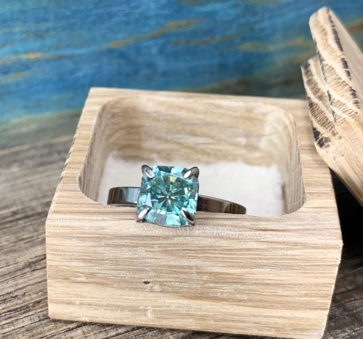A silver-toned ring with a cushion-cut blue-green stone, resting inside a light wooden box with a hinged lid.