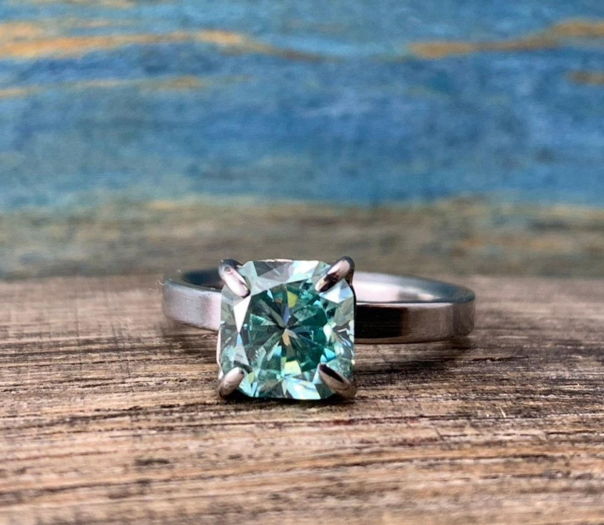 A silver-toned ring with a cushion-cut blue-green stone, positioned on a wooden surface with a blue and brown background.