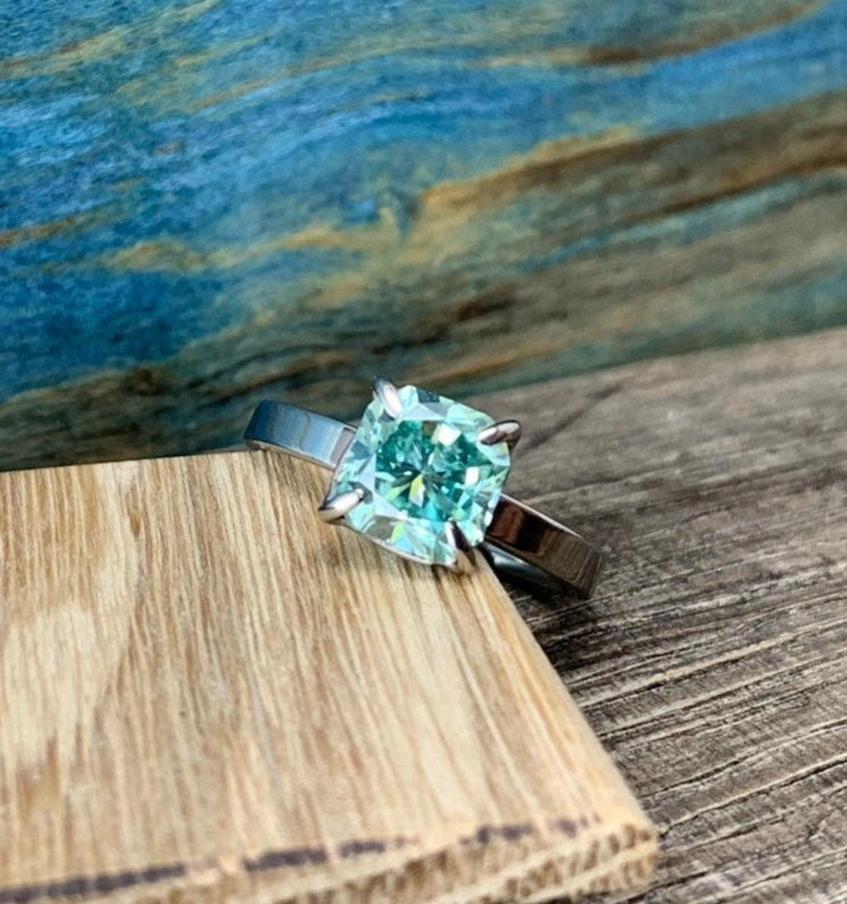A silver-toned ring with a cushion-cut blue-green stone, placed on a light wooden block against a blue and brown background.