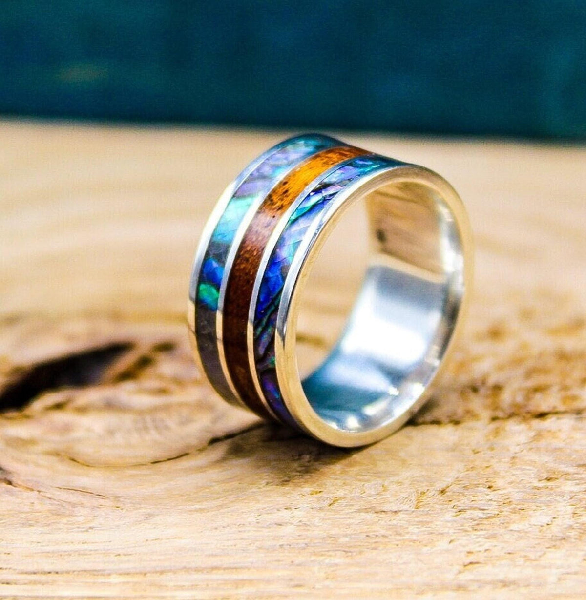A sterling silver ring resting upright on a wooden surface, featuring inlaid bands of iridescent blue abalone shell and brown mahogany wood.
